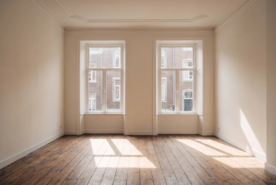 Een lege, frisgeschilderde kamer met eikenhouten vloer en twee hoge ramen, klaar voor een nieuwe huurder